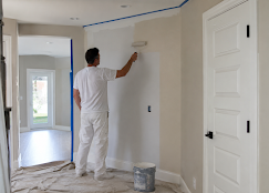 Painter rolling light-coloured paint on an interior wall with taped edges and a protected floor.