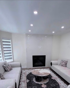 Light neutral living room with white sofas, recessed ceiling lights, and a dark fireplace.